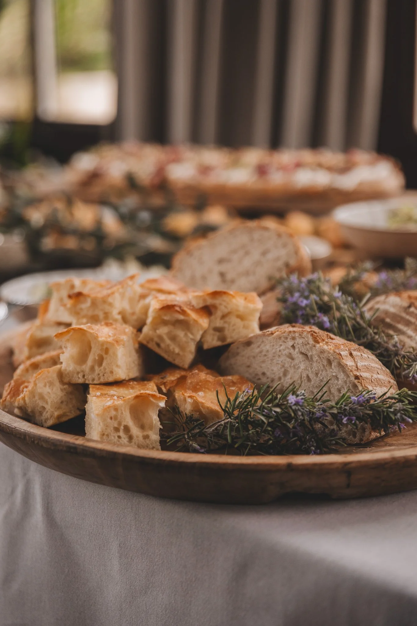 A wooden serving platter with various types of bread, including sliced bread and bread cubes, garnished with sprigs of lavender, on a white tablecloth.