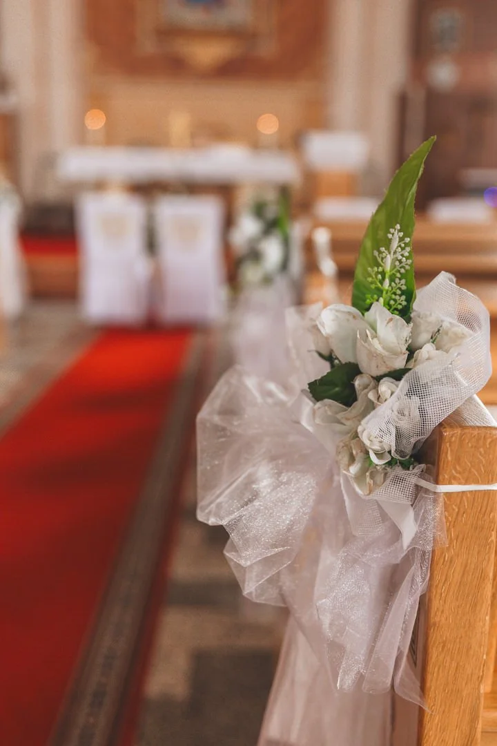 A small flower arrangement with white flowers, green leaves, and white mesh, tied to a wooden pew at a church, with an aisle and altar in the blurred background.