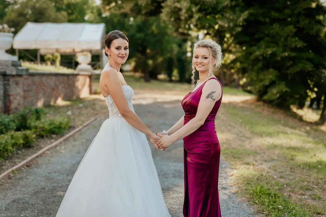 Two women, one in a white wedding gown and the other in a magenta dress, holding hands outdoors on a gravel path with trees in the background