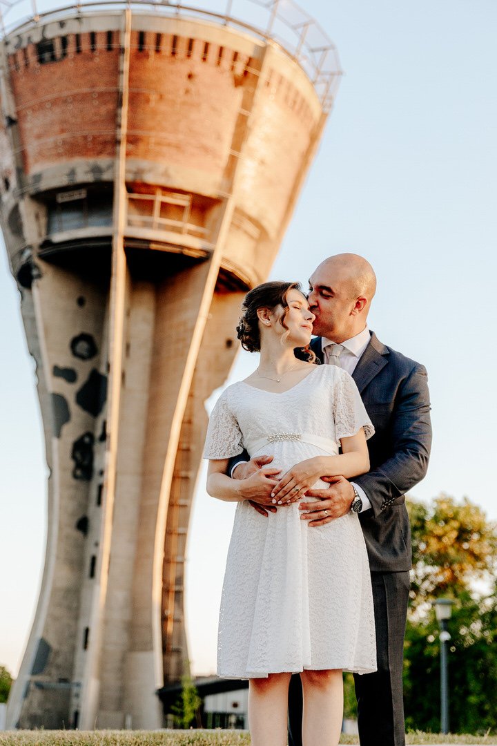 A couple embracing in front of a tall Vukovar water tower, curved concrete tower during golden hour, the man kissing the woman on her temple as she looks up with her eyes closed, wearing formal attire.