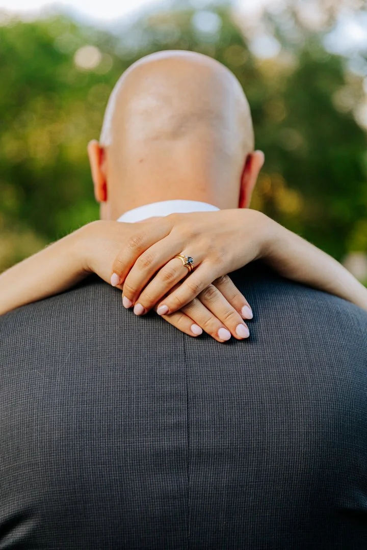A woman embraces a man from behind, resting her arms on his shoulders, with her hands with a wedding ring visible on his back. The man has a bald head and is wearing a gray suit. The background shows blurred green trees and sunlight.