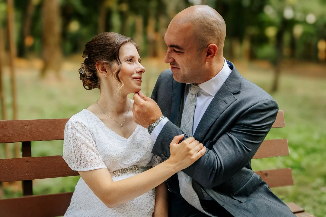 A couple sitting on a park bench, with the man gently holding the woman's chin, both smiling at each other lovingly.