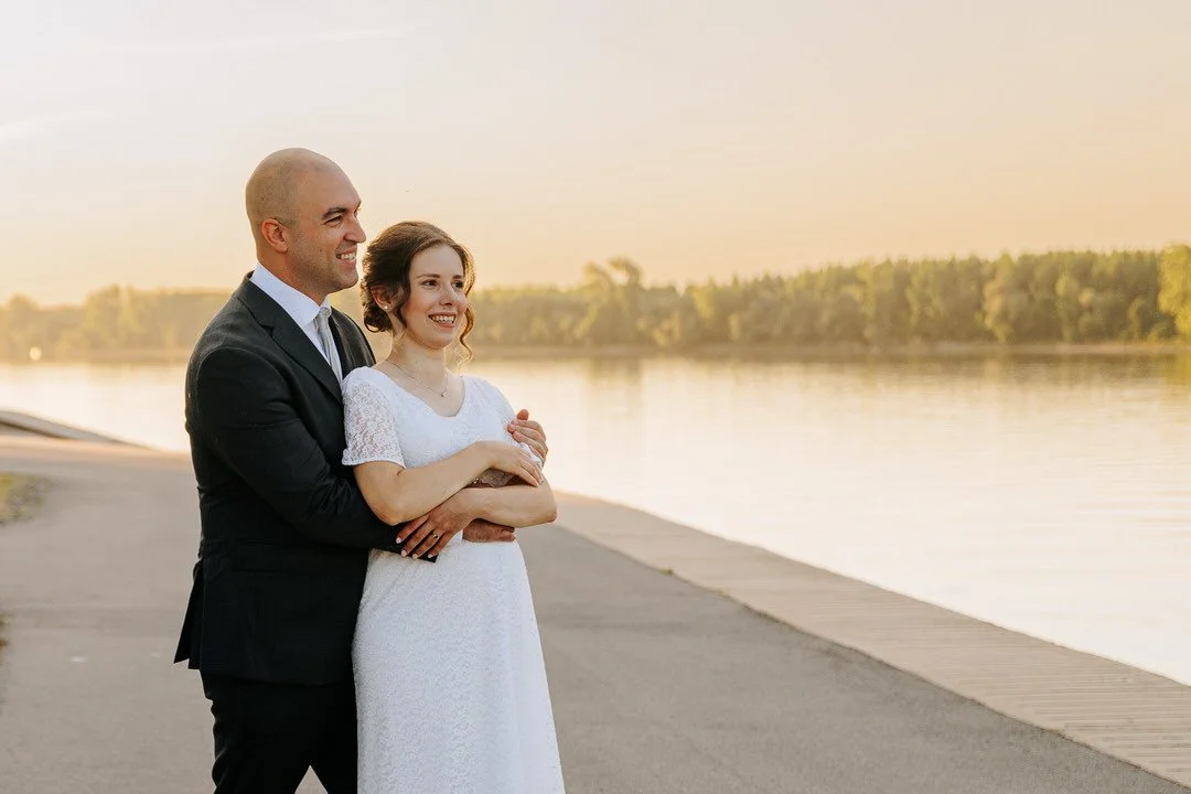 A ewlywed couple in formal attire standing by Dunav river at sunset, smiling and embracing each other.