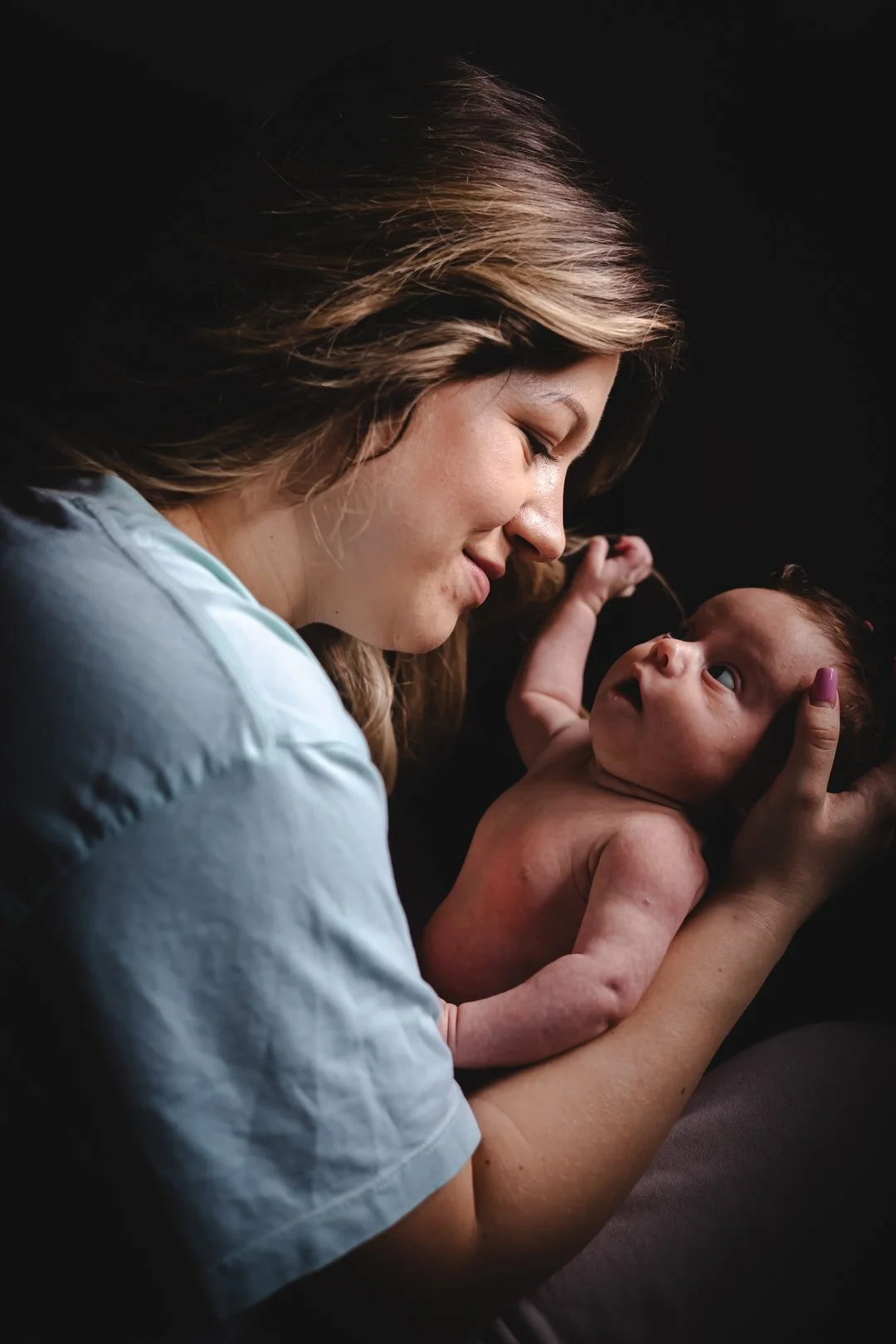 A mother holding a newborn baby in her hands, looking at the baby with a tender expression against a dark background.