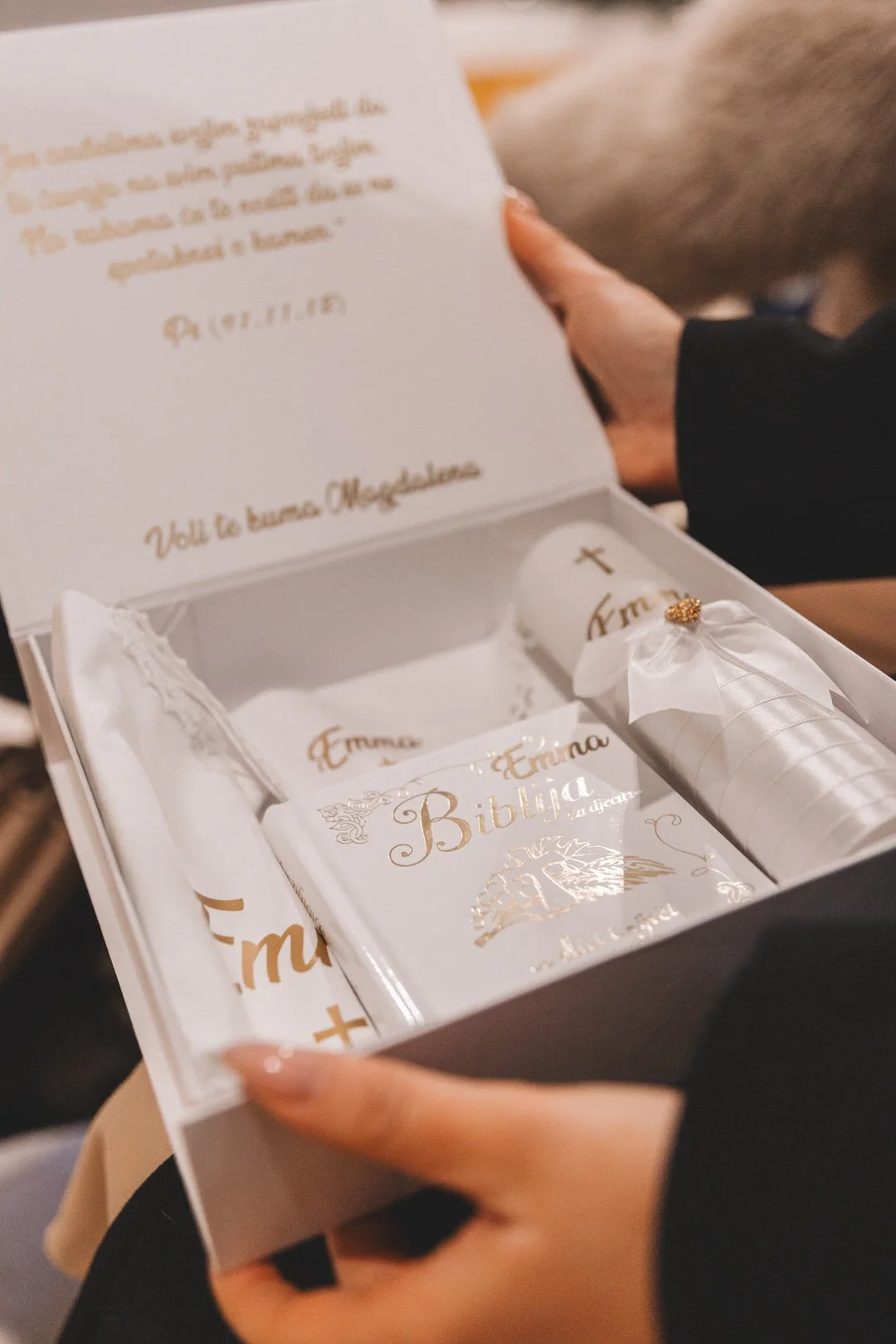 Open white box containing religious items, including a white candle, a wrapped Bible labeled 'Biblia', and a small wrapped item with a gold cross, with a hand visible holding the box.