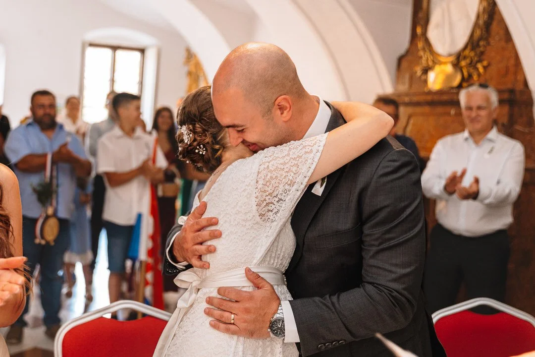 A couple embraces during a wedding ceremony, surrounded by guests in a decorated indoor venue.