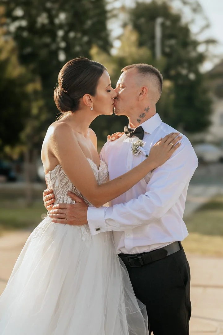 A bride and groom sharing a kiss outdoors, dressed in wedding attire, with trees and a blurred background.