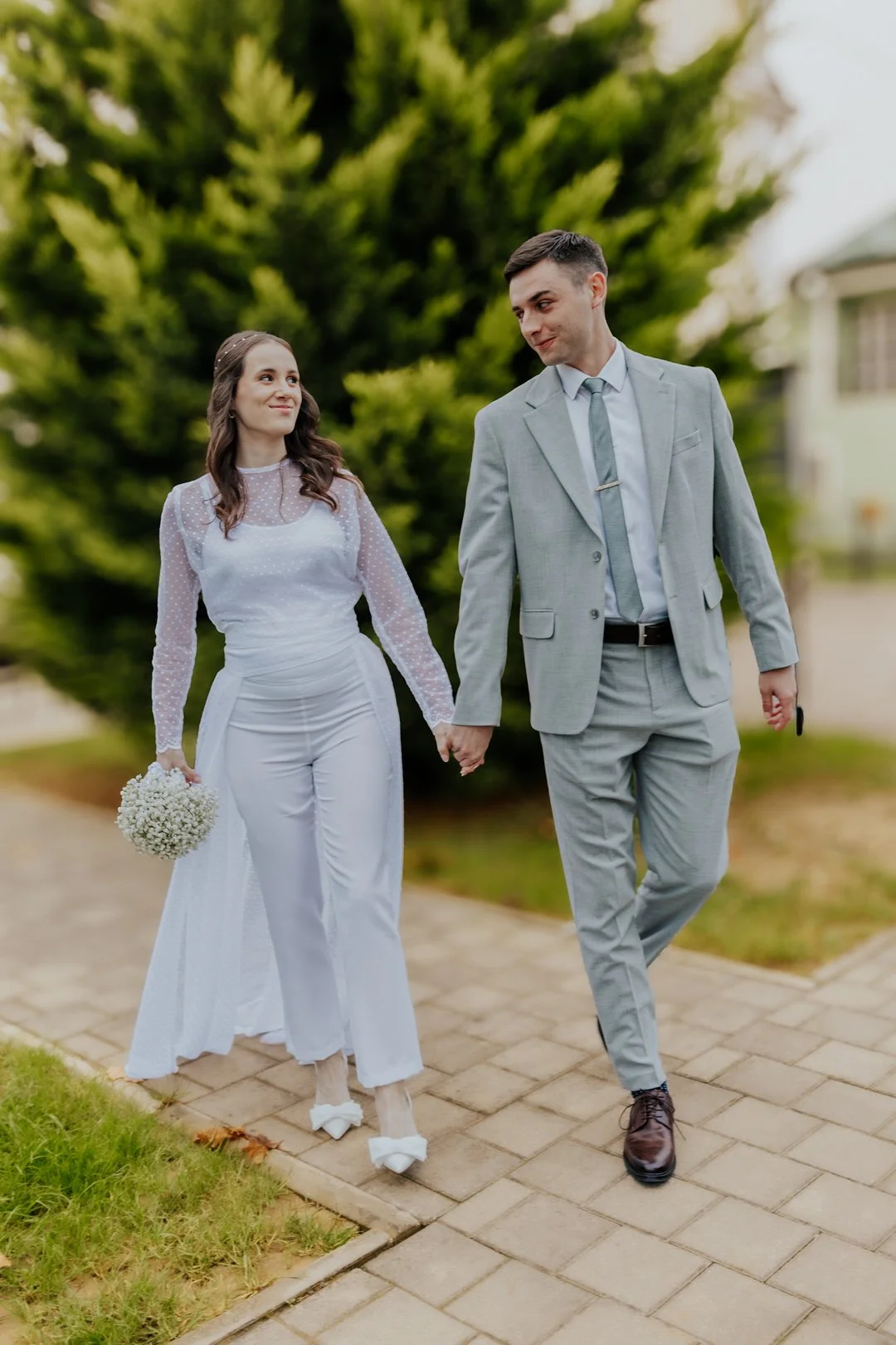 A bride and groom walking hand-in-hand outdoors, with the bride holding a bouquet, and a large green tree in the background.