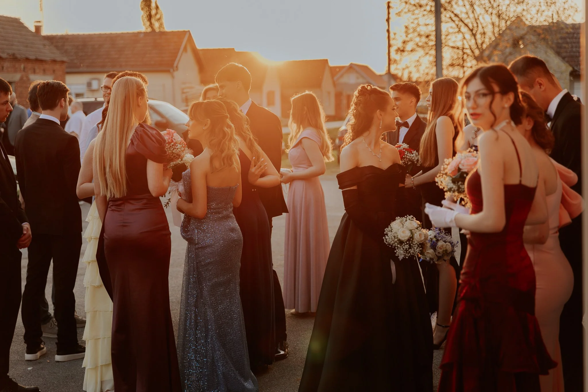 People dressed in formal attire, including women in evening gowns and men in tuxedos, gathered outdoors during sunset, holding flowers, likely attending a wedding
