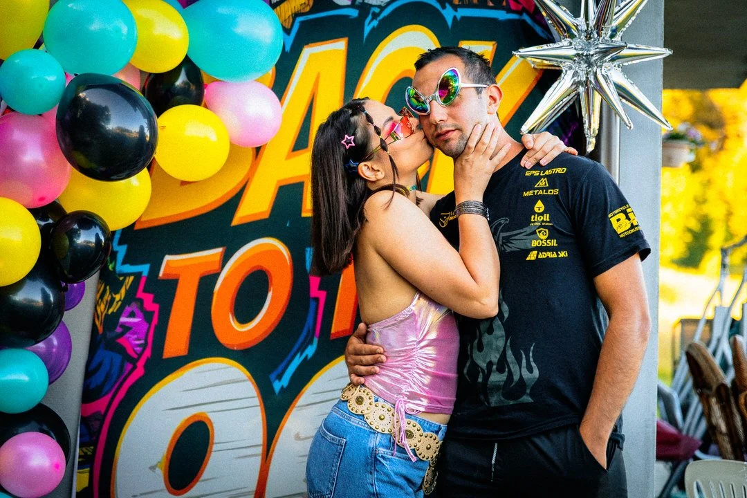 A couple wearing colorful sunglasses, hugging, and kissing at a party with balloons and a vibrant backdrop with "back to 90's" text 