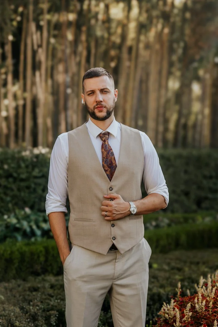 A man wearing a beige vest, white shirt, paisley tie, and beige pants standing outdoors in a garden with trees in the background.