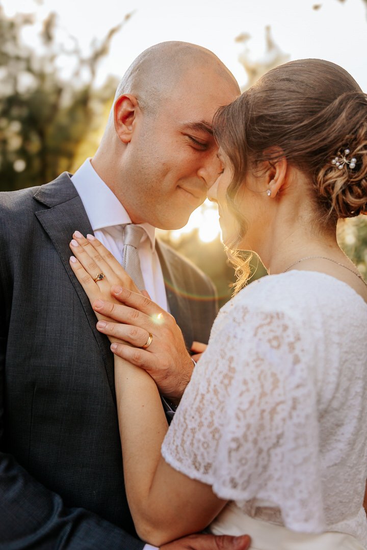 A bride and groom embrace outdoors during sunset, touching foreheads with eyes closed, both wearing wedding attire, with the bride in a lace dress and the groom in a suit, capturing a tender moment in park forest Adica in Vukovar