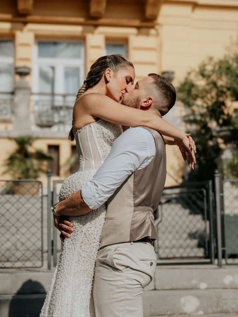 A couple sharing a kiss outdoors, with the woman in a white dress and the man in a light-colored shirt and vest.