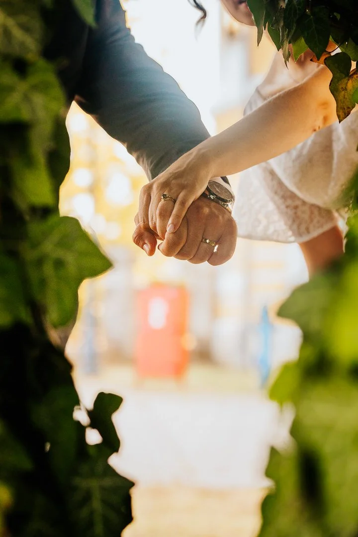 Close-up of a man and woman holding hands with wedding rings, surrounded by green foliage, in an outdoor setting with blurred background.