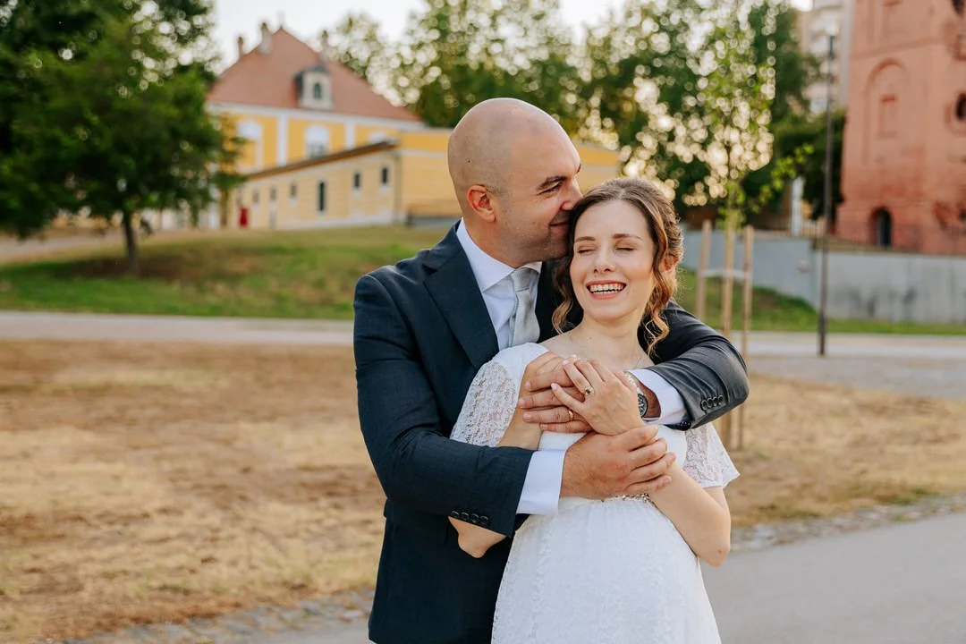A man in a suit embraces a woman in a white dress, kissing her on the head while she smiles with eyes closed outdoors.