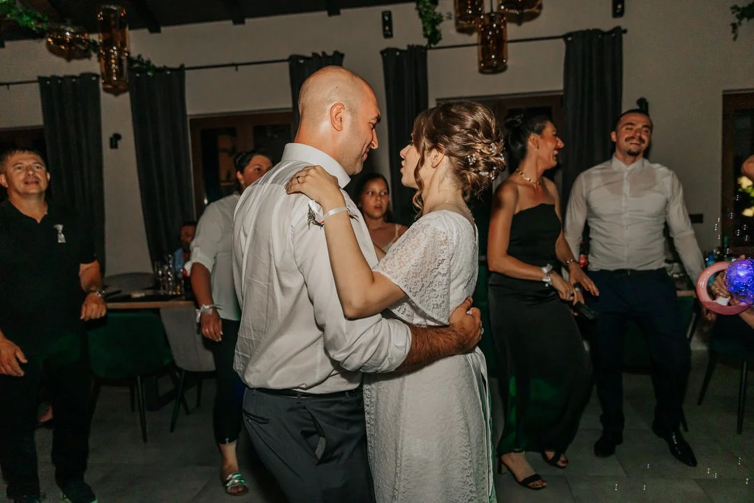 A wedding reception with a couple dancing in the center, surrounded by guests who are smiling and enjoying the celebration. The bride is wearing a white lace dress, and the groom is in a white shirt. The venue has dark curtains and elegant lighting.