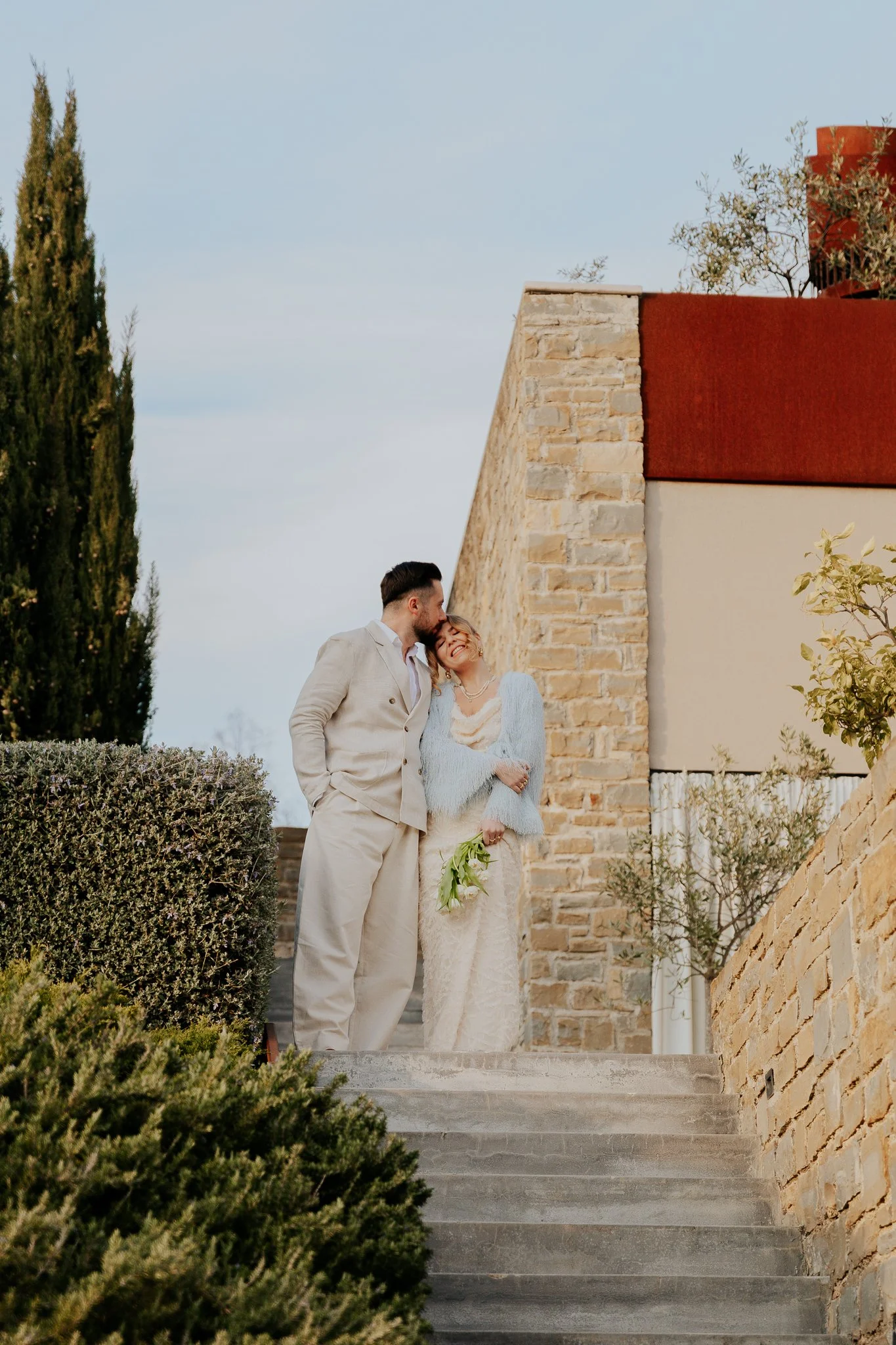 A couple in wedding attire stands on outdoor steps, embracing and smiling. The man is wearing a light gray suit, and the woman is in a lace wedding dress holding a bouquet. Behind them is a stone building with a red roof, and lush greenery surrounds 