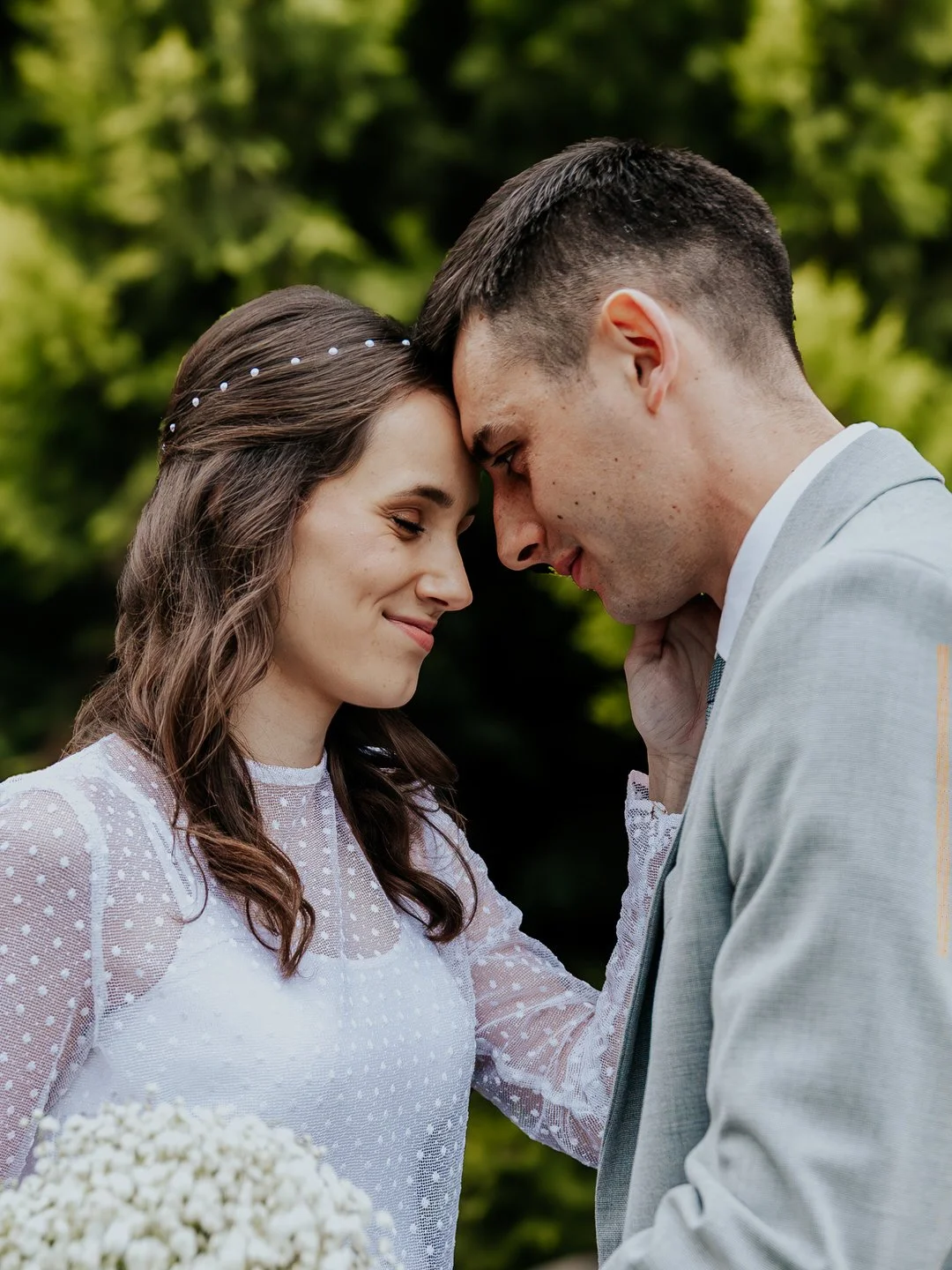 A bride and groom with foreheads touching, eyes closed, sharing a tender moment outdoors. The bride wears a white lace dress with her hair in soft waves and a pearl headband, holding a bouquet of white flowers. The groom wears a light gray suit with 