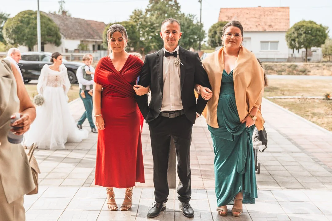 Three people walking arm in arm outdoors at a formal event or wedding. The person in the center is a man in a tuxedo, flanked by two women in elegant dresses, one in red and one in teal.