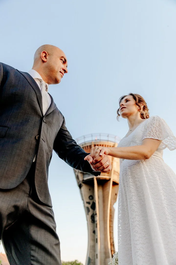A couple holding hands, with the man dressed in a suit and the woman in a white dress, standing outdoors near a Vukovar water tower under a clear sky.