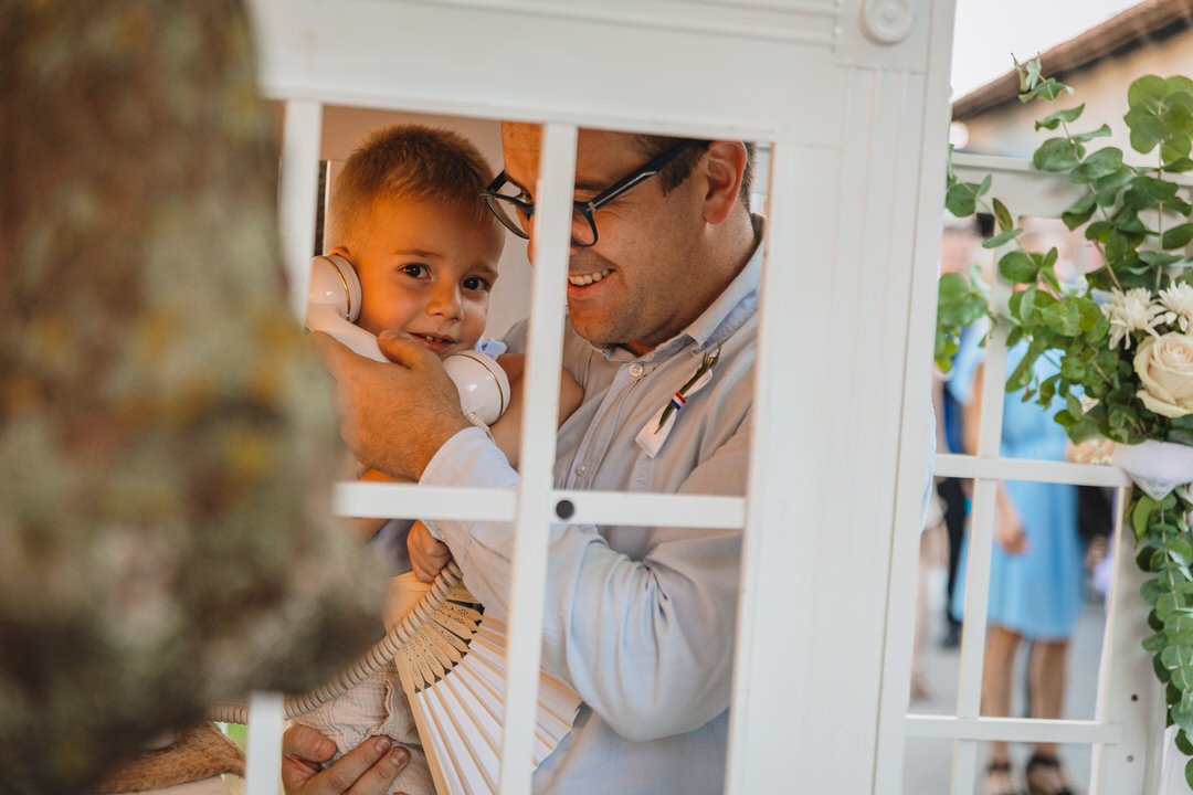 A man and a young boy are seen through a birdcage, smiling and embracing. The boy is holding a vintage telephone receiver to his ear, with a tree trunk partially visible in the foreground and flowers in the background.