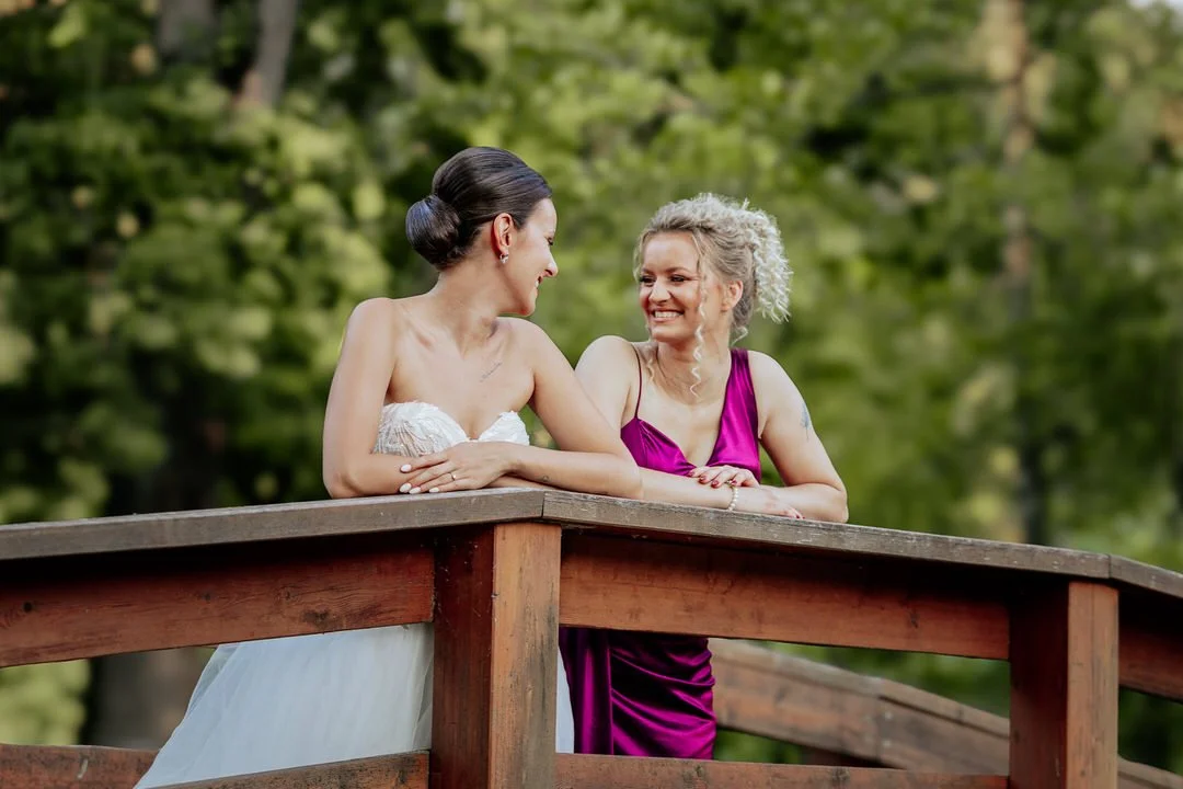 Two women are smiling and talking while leaning on a wooden railing outdoors surrounded by green trees. One woman is wearing a white strapless dress, and the other is wearing a magenta dress.