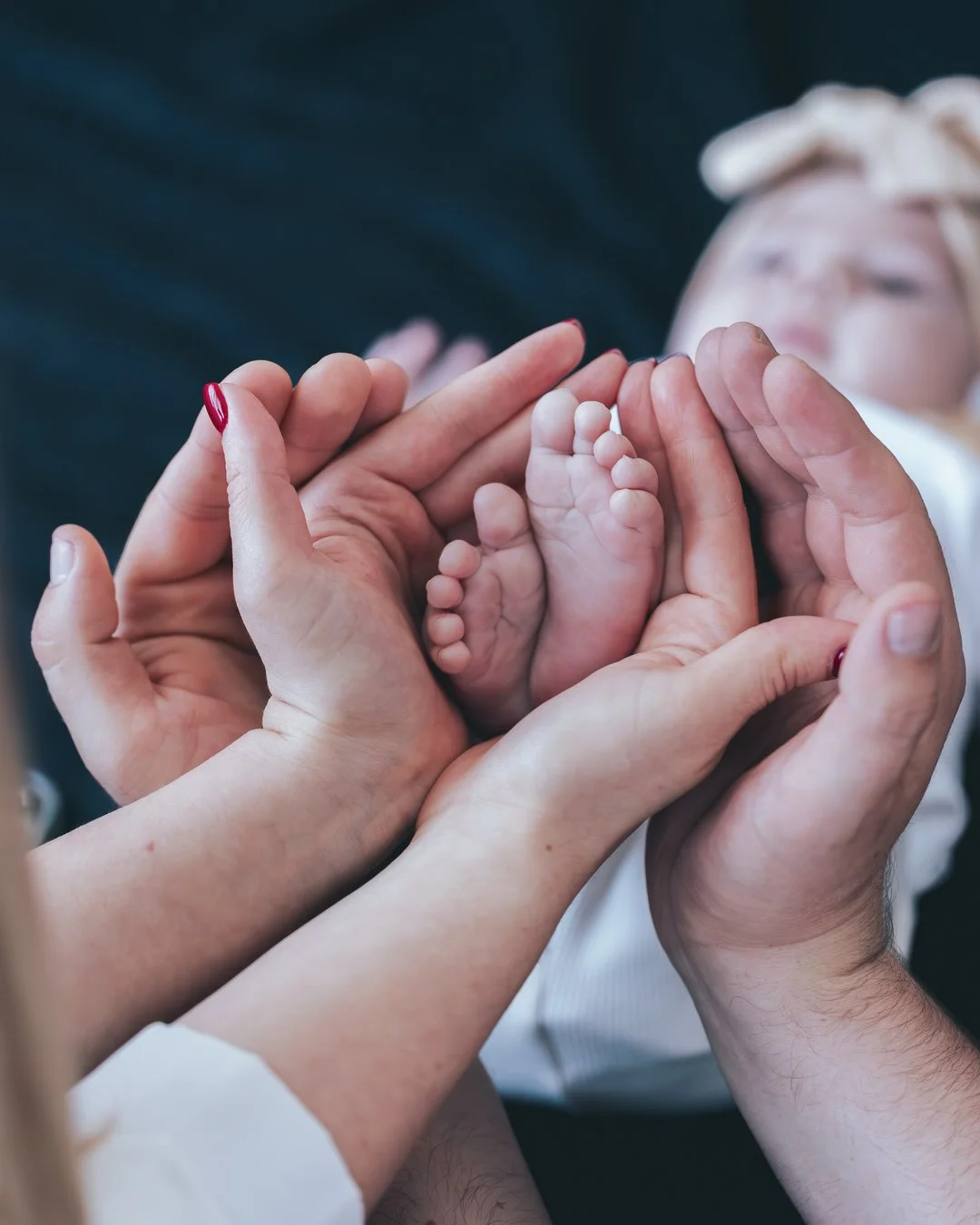 mother and father hands gently cradle tiny baby feet, with a blurred background of a person in a white outfit.