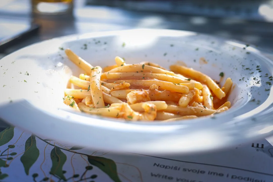 A white bowl of spaghetti with sauce, garnished with herbs, on a table outdoors with sunlight shining.