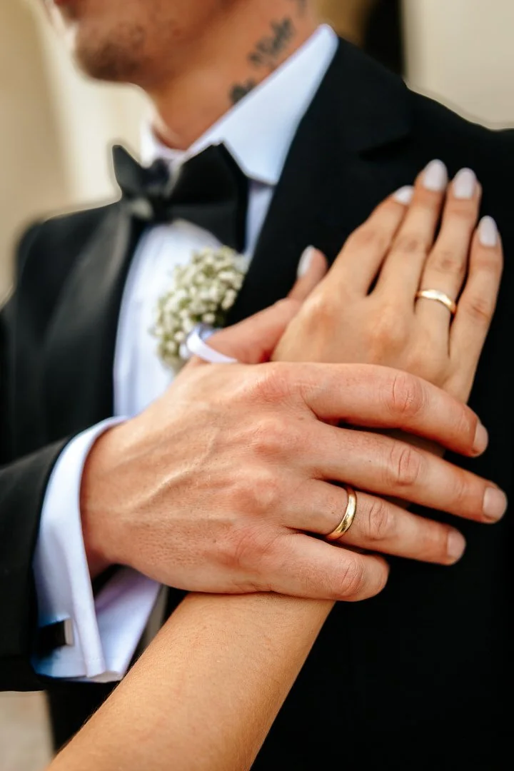 Close-up of a couple's hands with wedding rings, the man's hand on the woman's arm, wearing formal wedding attire, including a tuxedo and boutonniere.