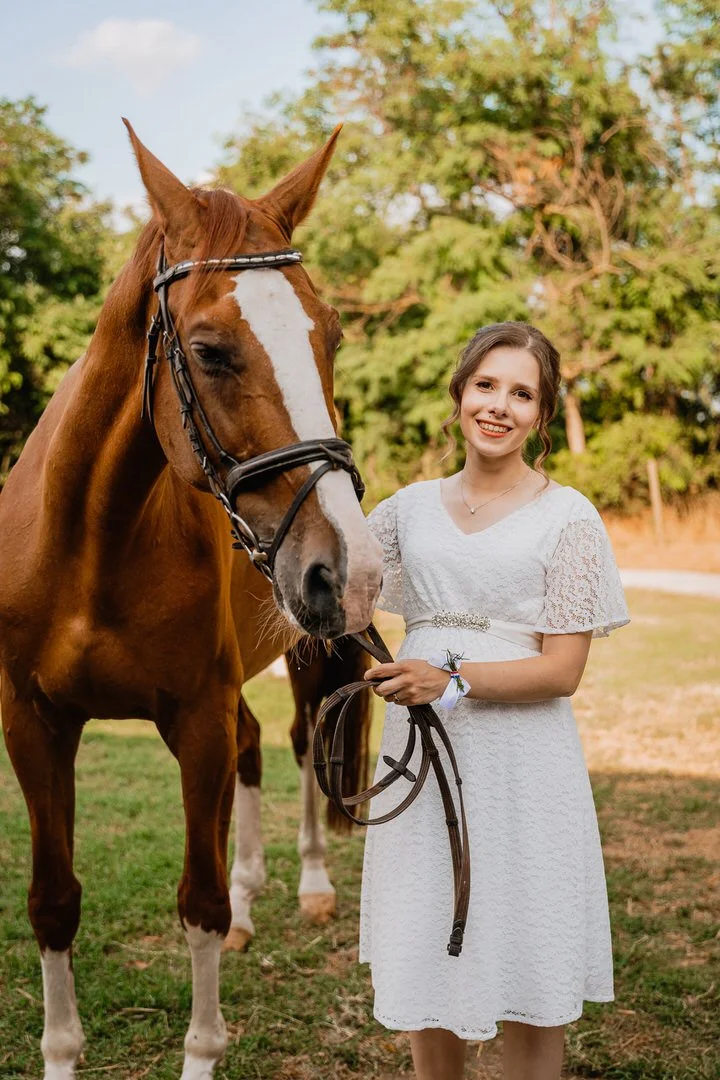 A bride in a white dress standing next to a brown horse with a white stripe on its face outdoors in Dunavski Raj venue 