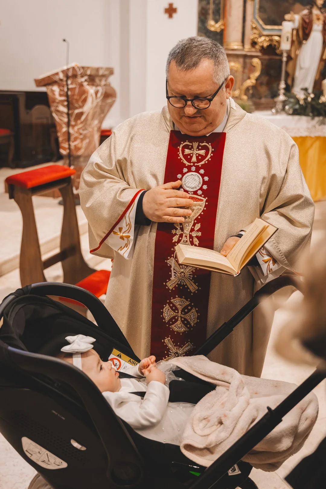 A priest in ornate vestments is praying with his hand over a baby's stroller during a Croatia baptism in Slavonija, inside a church while reading from an open book.