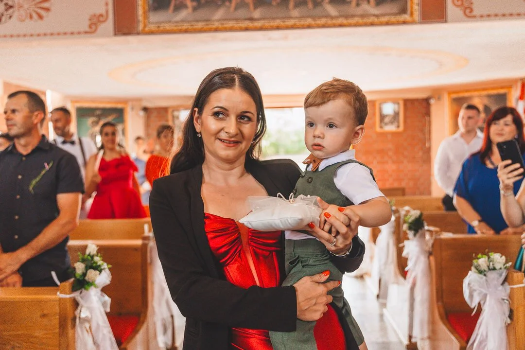 A woman holding a young boy at a church ceremony, with guests standing in pews decorated with white ribbons and flowers.
