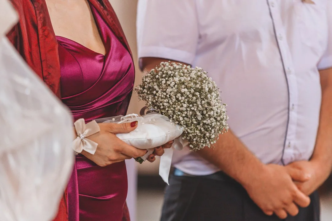 Person holding a bouquet of white flowers and a satin pillow, with another person wearing a white shirt nearby.