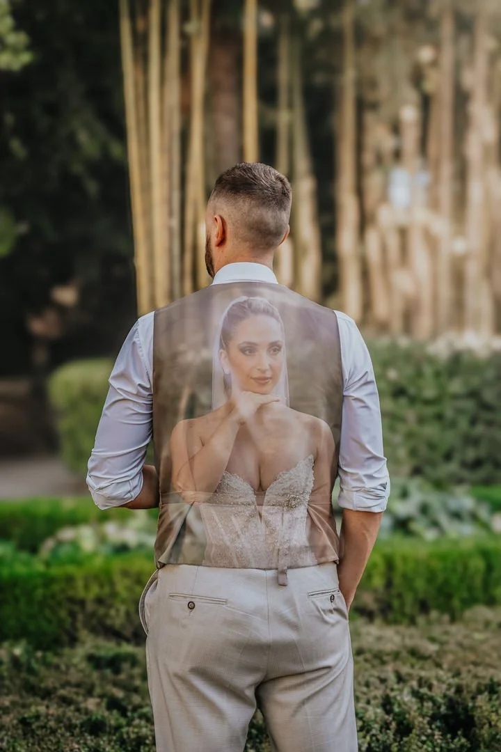 A man in dress pants and a shirt stands outdoors with his back to the camera, wearing a vest with a picture of a woman in a wedding dress printed on it, appearing to be looking at the picture.