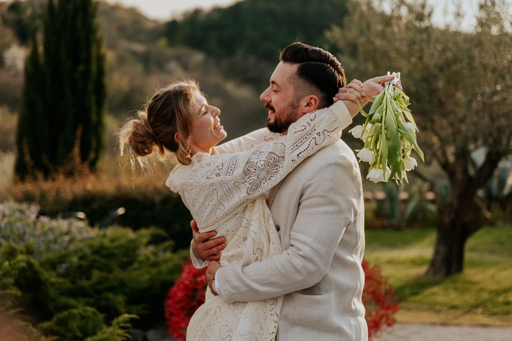 A man and woman sharing a joyful moment outdoors, with the woman holding a bouquet of white tulips. The woman is wearing a cream-colored lace dress, and the man is dressed in a light-colored suit. They are smiling and looking at each other, surrounde