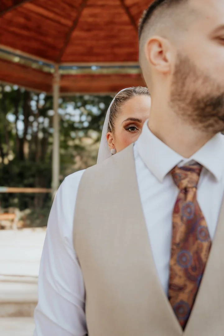 A bride peeking from behind a groom at an outdoor wedding, with trees and a wooden canopy in the background.