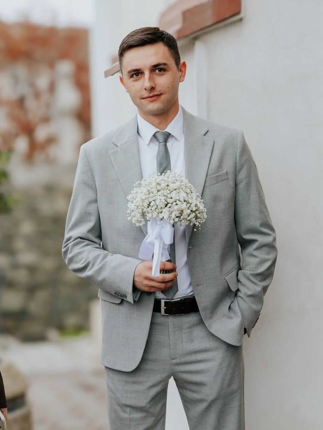 Groom in a light gray suit holding a bouquet of white flowers, standing outdoors by a white wall with a blurred background.