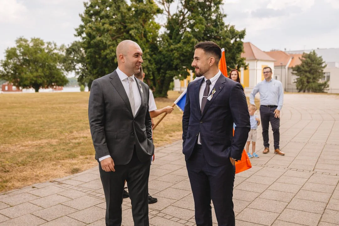 Two men in suits talking outdoors in front of Eltz Castle in Vukovar, Croatia, with wedding guests, children, and flag bearers in the background.