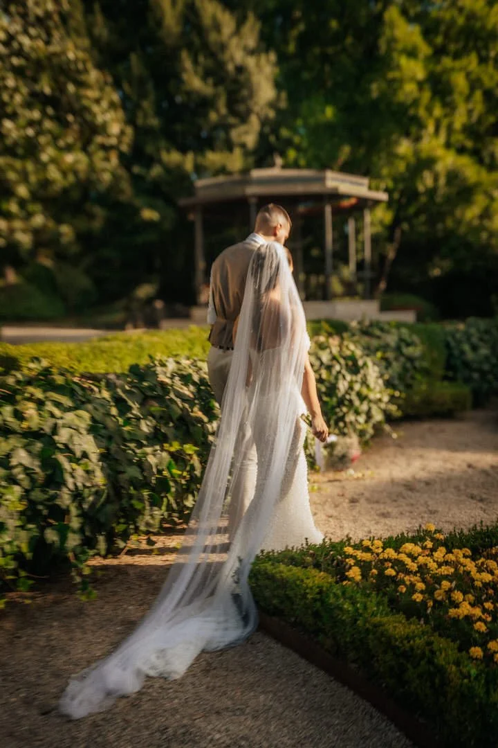 Bride and groom walking together through Park Angiolina in Opatija, Kvarner, during sunset, surrounded by lush greenery and yellow flowers.