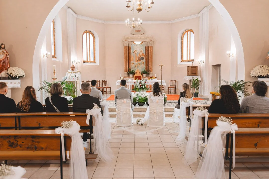 A small church with a wedding ceremony taking place. The altar is decorated with flowers and religious icons, with a priest officiating the wedding. The bride and groom are seated, facing the altar, with guests sitting on wooden pews decorated with w