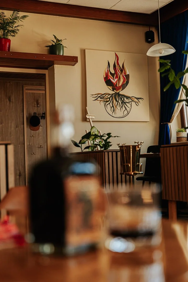 Interior of a cozy room with a modern wooden decor, a large abstract painting of a flame with roots on the wall, plants, a table setting with a wine glass and a bottle, and a hanging light fixture.