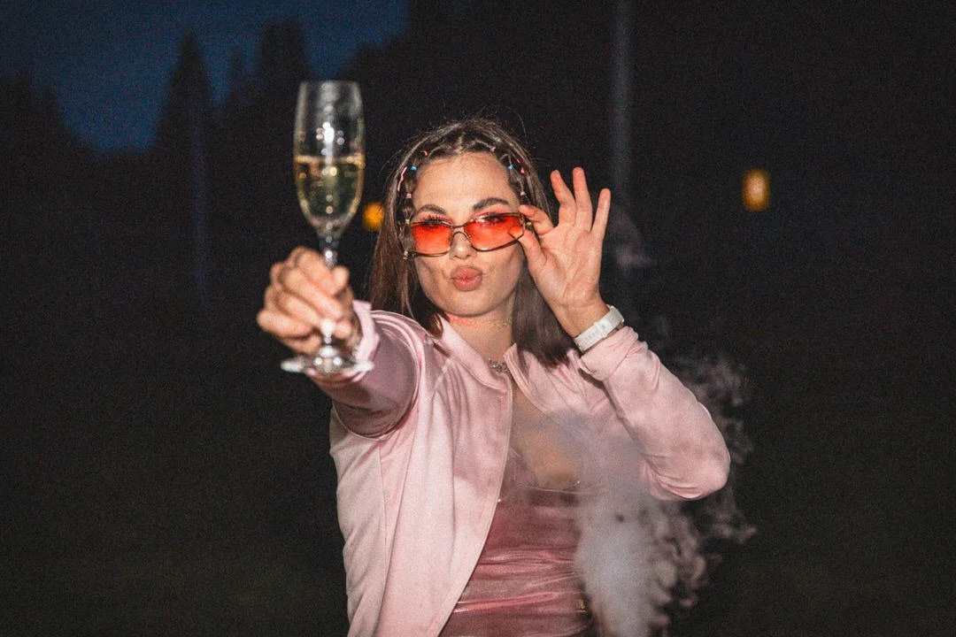 A woman in pink sunglasses and a pink satin jacket holding a glass of champagne at a birthday party.