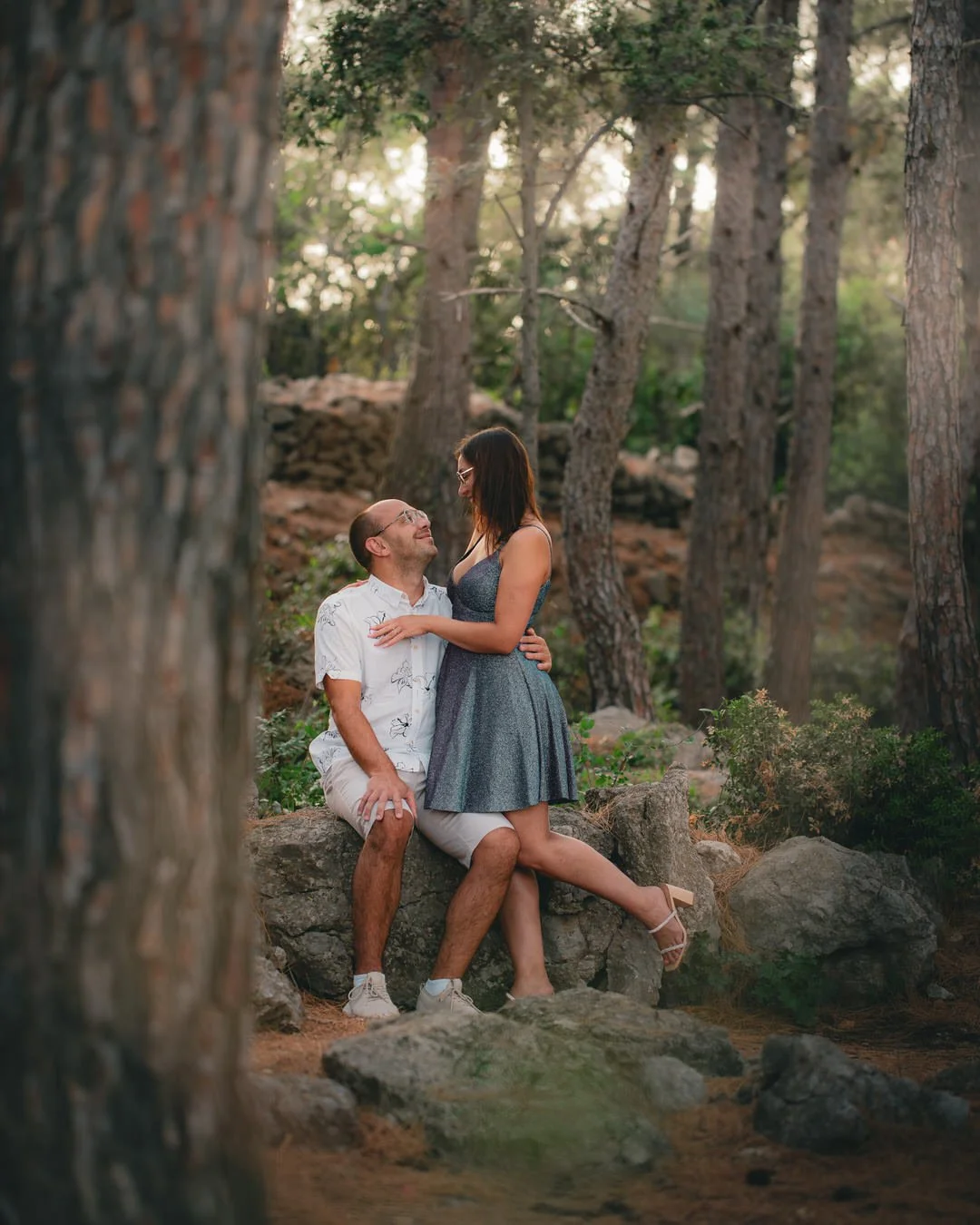 A couple sitting on rocks in a wooded area, the man kneeling on one knee and the woman standing close to him, both smiling and looking at each other, surrounded by trees with sunlight filtering through at Plav Beach on Krk Island, Croatia