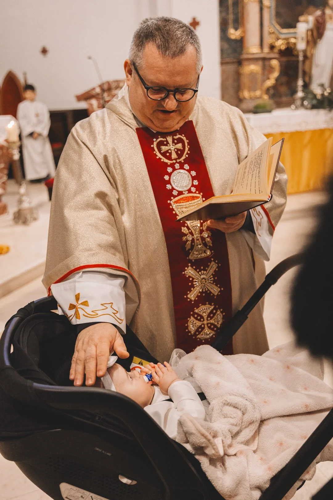 A priest in ornate vestments conducts a Croatia baptism ceremony in Slavonija at a church altar, standing beside a baby in a car seat who is looking up  and wrapped in a blanket.
