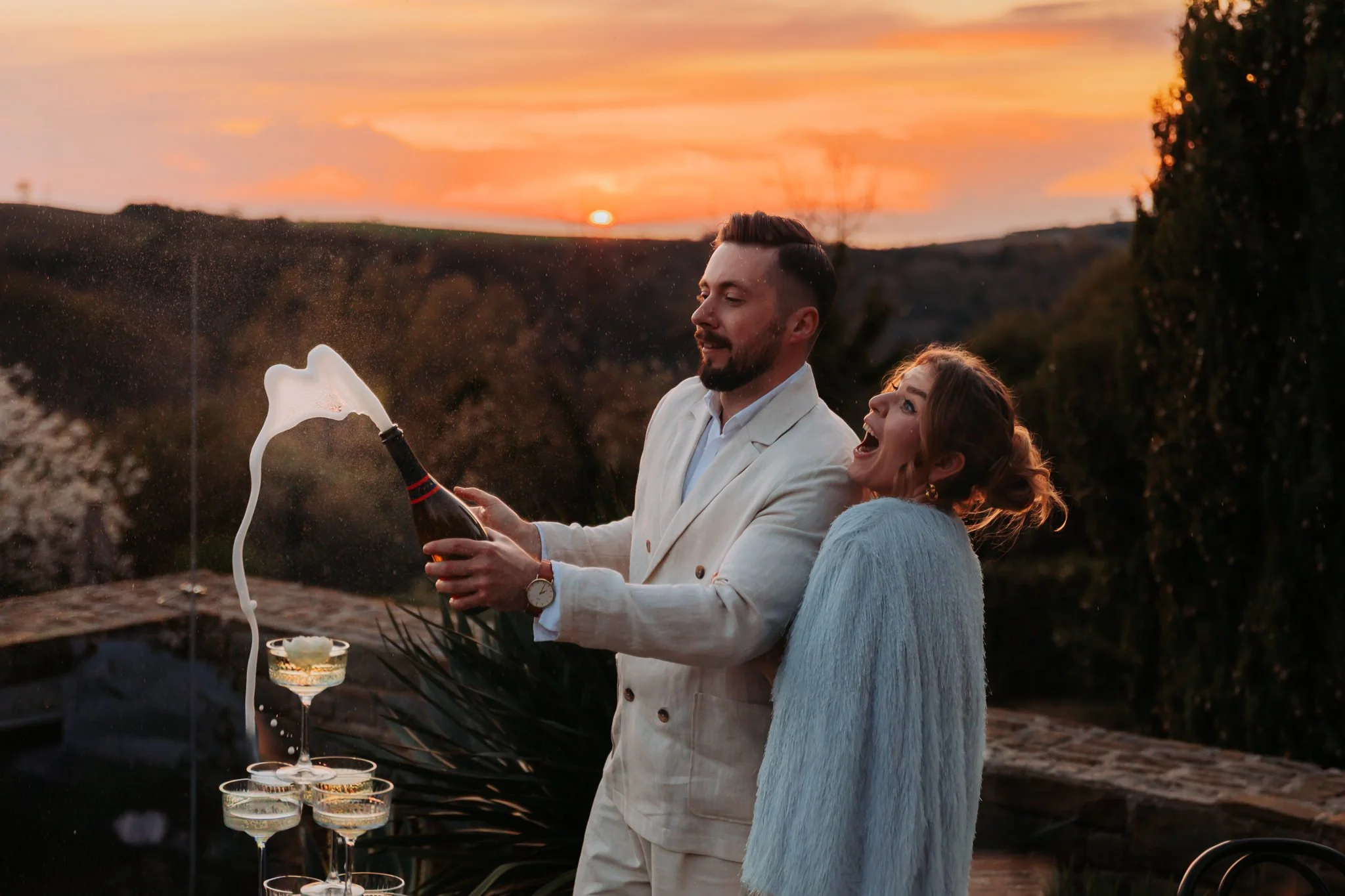 A man and woman celebrating wedding outdoors at sunset in San Canzian Istria,Croatia , with the man opening a bottle of champagne and the woman smiling excitedly. There are glasses of champagne and a tiered tray with more glasses on a table.