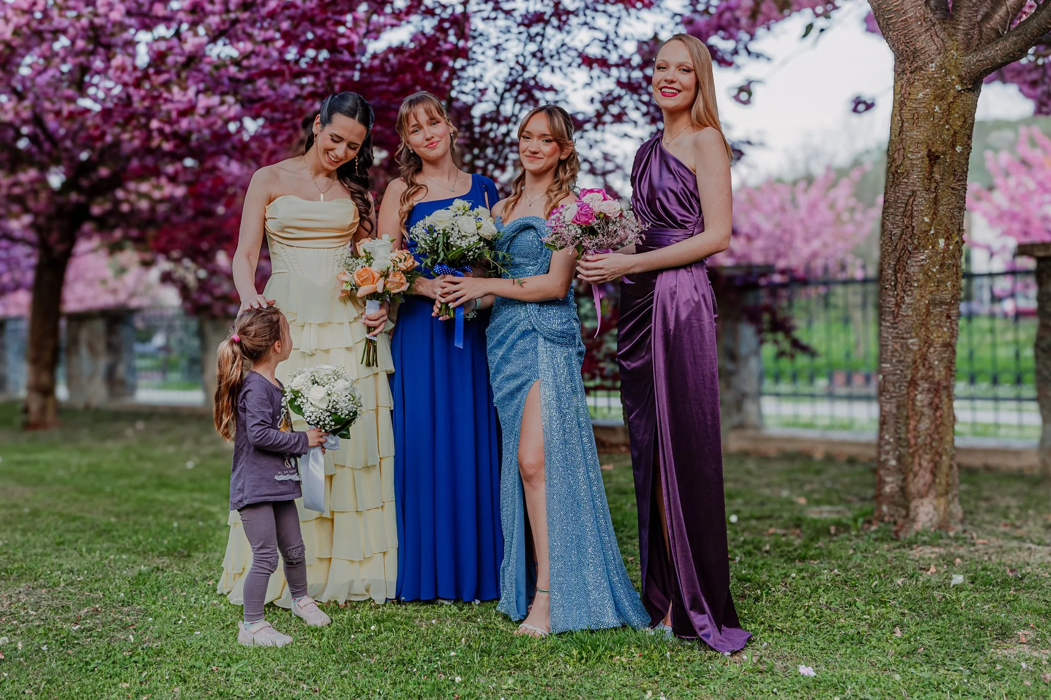 Four women in colorful dresses holding bouquets of flowers standing outside among pink blossoming trees; one young girl in purple holding a small bouquet, looking up at the women.