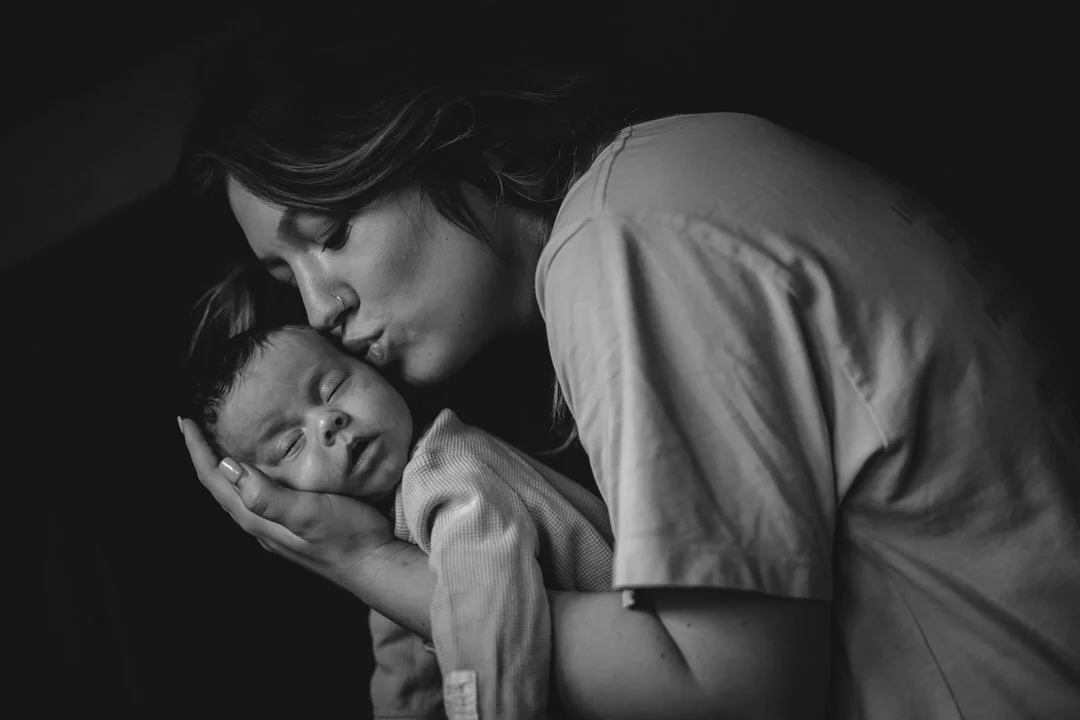 A mother tenderly holding a sleeping baby, kissing the baby's forehead in a black and white photograph.