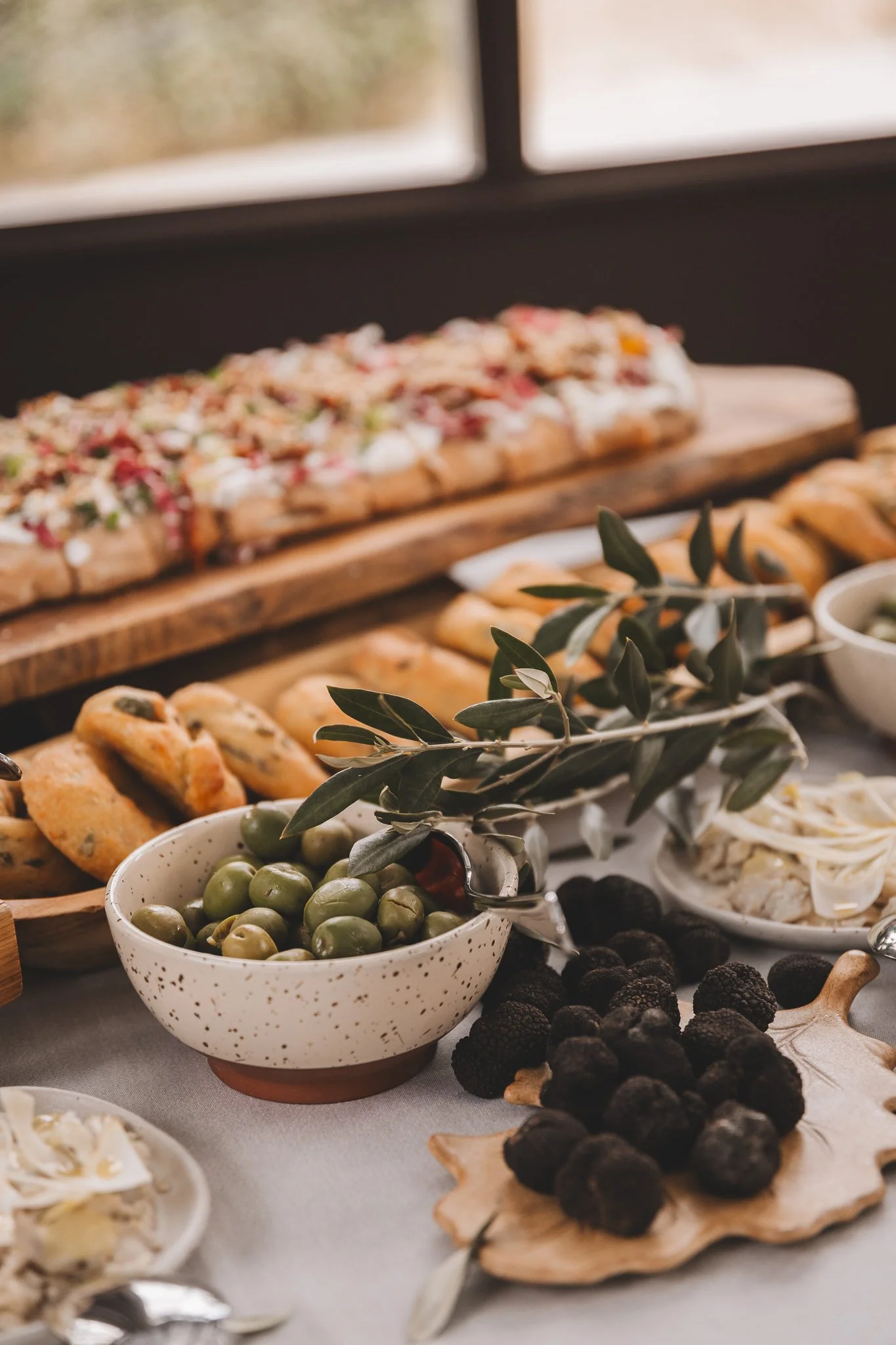 A variety of appetizers and snacks on a table, including a bowl of green olives, truffles, bread rolls, and a spread with herbs and toppings, set against a blurred background with a window.