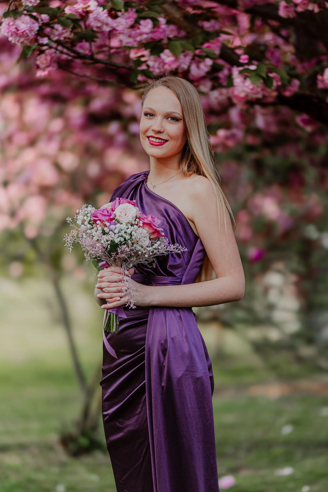 A young woman in a purple satin dress holding a bouquet of pink and white flowers, standing outdoors with pink blossoms in the background at the park in Orahovica Croatia