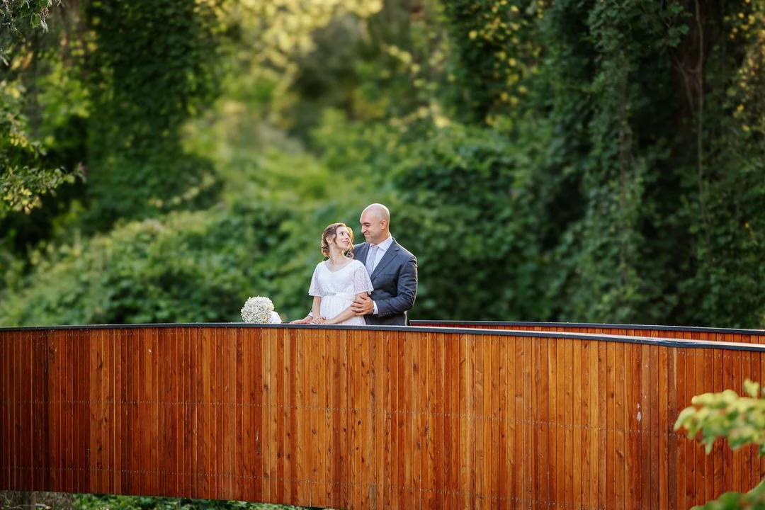 A couple standing on a wooden bridge in park forest Adica in Vukovar, surrounded by lush green trees, smiling at each other, with a bouquet of flowers on the bridge.